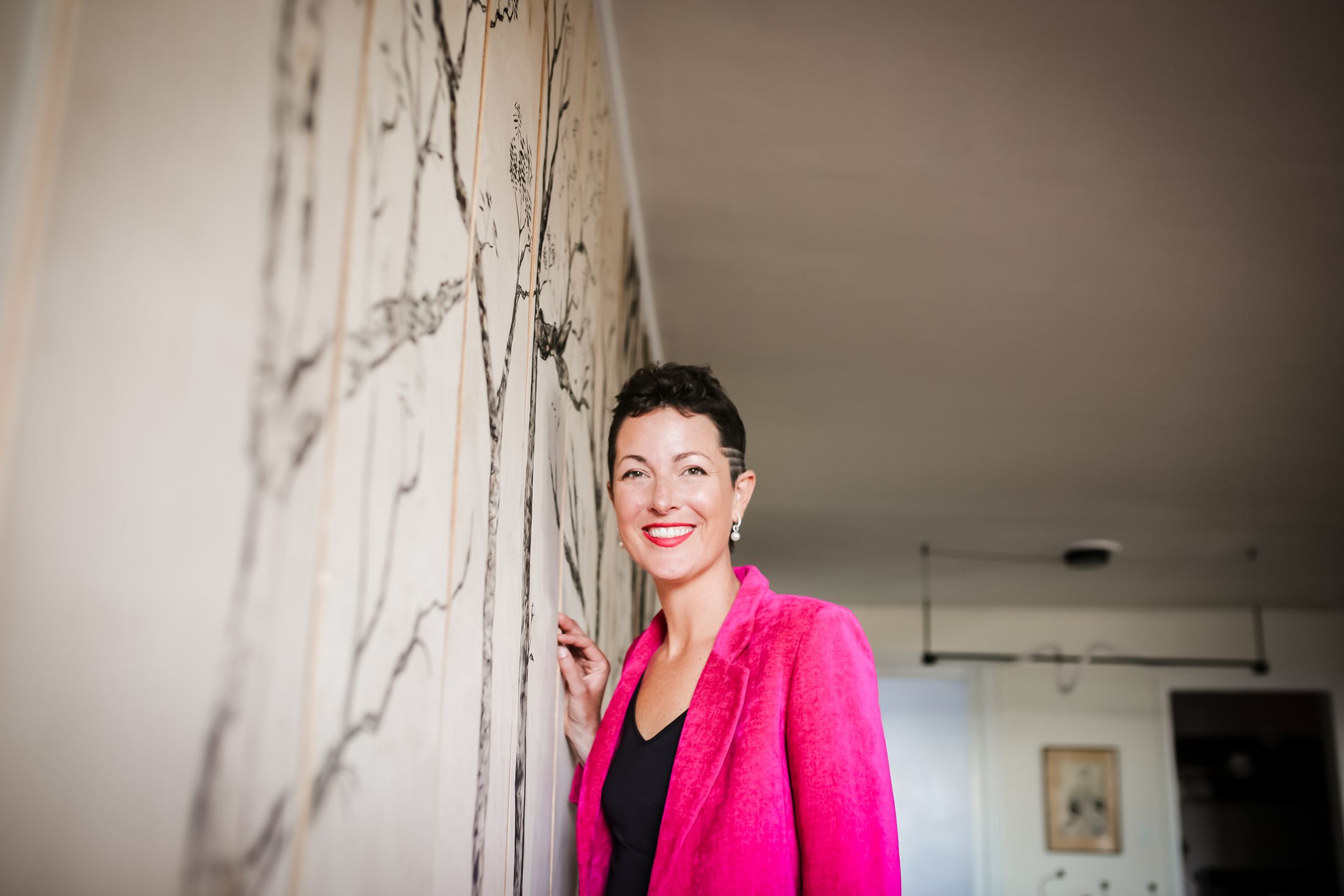 Louise laughing in a pink suit beside her hand-painted tree wall