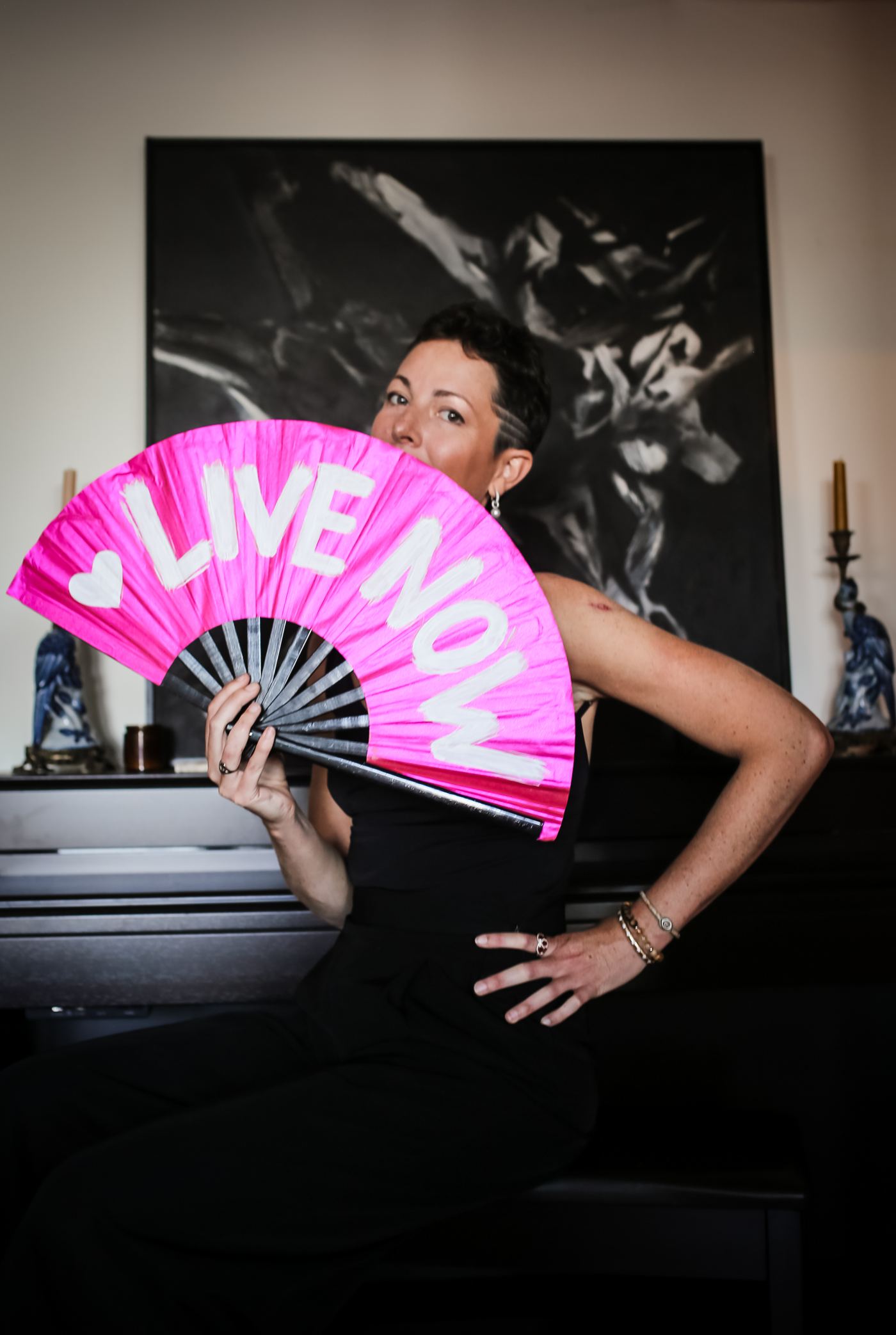 Louise glancing over a pink LIVE NOW fan, black-and-white artwork behind her